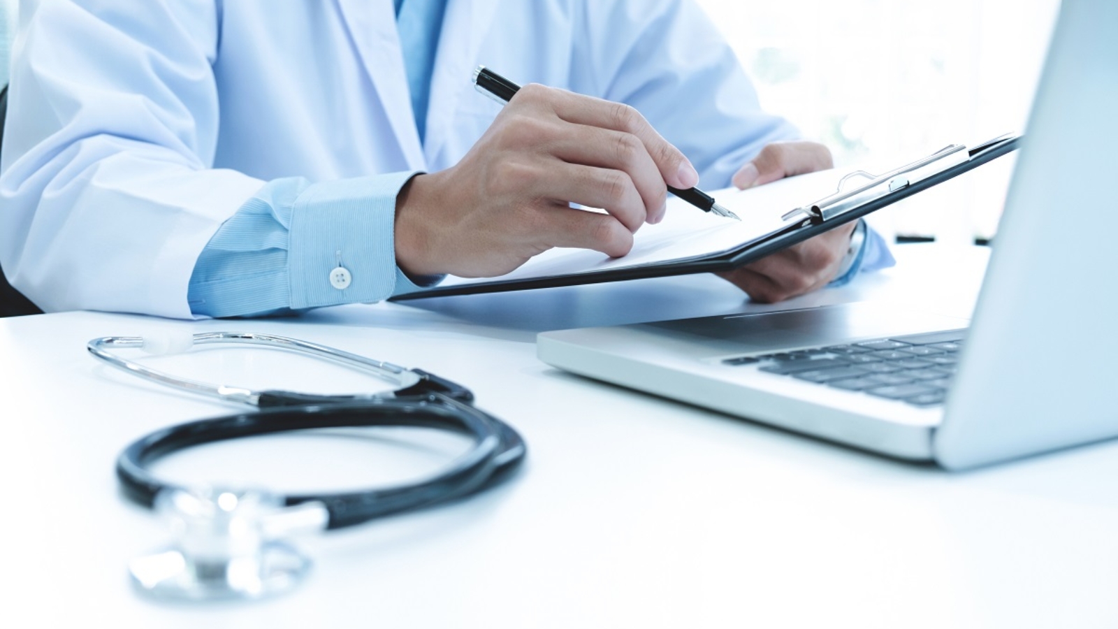 Doctor working with laptop computer and writing on paperwork. Hospital background.